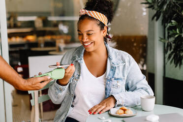 Cheerful young woman paying with her phone at a coffee shop in a contactless manner, showcasing electronic banking convenience. Female customer using NFC technology to settle her bill in a cafe. - JLPSF30717