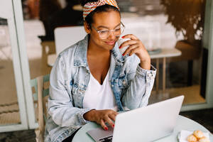 Eine Content-Autorin, die in einem Café arbeitet, lächelt, trinkt Kaffee und schreibt an ihrem Laptop. Eine selbstbewusste junge Frau, die die Freiheit des modernen digitalen Nomadentums genießt. - JLPSF30718