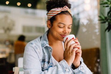 Junge Frau in einem gemütlichen Café, die eine Tasse Kaffee genießt. Ihr lächelndes Gesicht spiegelt Glück wider, als sie sich einen Moment Zeit nimmt, um sich bei diesem heiteren Solo-Date zu entspannen. - JLPSF30720