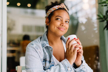 Pensive young woman enjoys a hot beverage in a coffee shop, exuding authentic happiness in this solo date moment. Young woman enjoying a moment to herself in a cozy cafe. - JLPSF30721