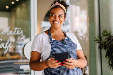 Happy female entrepreneur in a small café holding a digital tablet, engaging customers with a friendly smile. She embodies success in business with wireless technology in a bustling coffee shop setting. - JLPSF30724