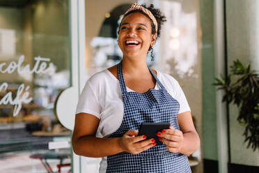 Happy young entrepreneur embraces technology in a vibrant café, holding a digital tablet in hand. Female cafe owner stands confidently in her successful small business. - JLPSF30725