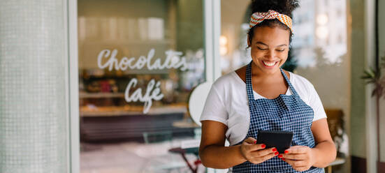 Happy female entrepreneur standing in a coffee shop, using a digital tablet for easy cafe management. Successful small business owner reading her online reviews, staying connected to her customers. - JLPSF30726