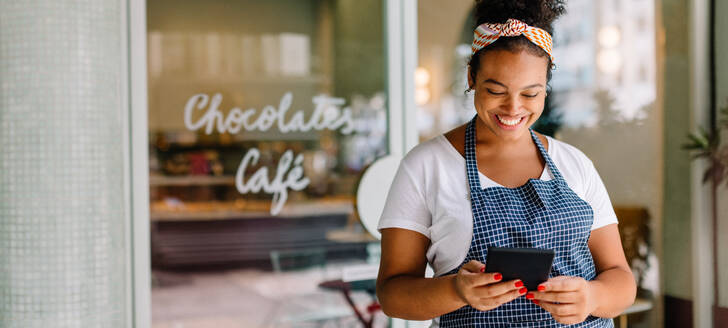 Happy female entrepreneur standing in a coffee shop, using a digital tablet for easy cafe management. Successful small business owner reading her online reviews, staying connected to her customers. - JLPSF30726