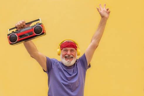 Cheerful elderly man in red head band and headphones holding cassette player with excited face expression while standing against yellow background - ADSF45764