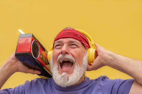 From below elderly bearded male in casual clothes with headband and headphones with cassette player screaming while listening to music - ADSF45766