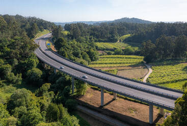 Aerial view of cars driving on asphalt road with lanes and bridge with pillars constructed on valley with agriculture fields while going through green fields in countryside - ADSF45783