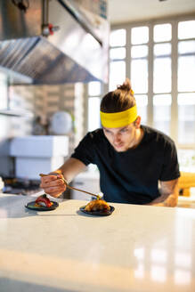 Concentrated young male with yellow headband looking down and serving tuna fish sushi in plate with ingredients at kitchen counter against daylight - ADSF45791