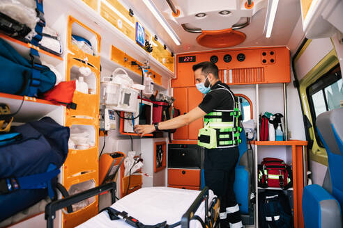 Side view of concentrated guy in uniform and medical mask checking facility in automobile while working as paramedic - ADSF45794