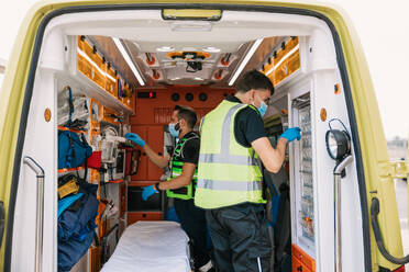 Side view of busy emergency medical technicians in uniform and protective masks checking equipment in car before responding to calls - ADSF45795