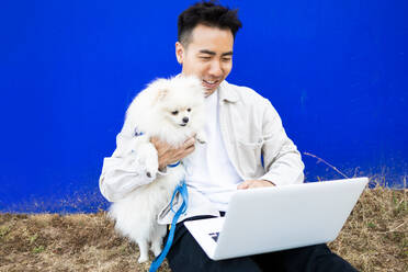 Happy Asian male dog owner in casual wear with cute Pomeranian Spitz and computer video calling while relaxing on weekend - ADSF45798