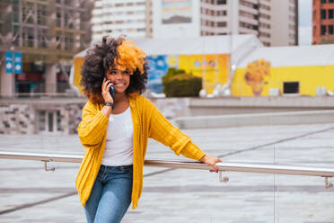 Positive black female with yellow and black afro hair in casual clothes standing on city street by modern apartment while making phone call and looking away - ADSF45804