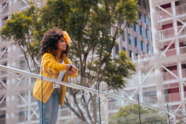 Low angle of positive young African American female with yellow and black afro hair in casual clothes leaning on glass railing against modern city building while looking away - ADSF45805