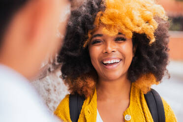 Back view of young anonymous man and cheerful woman with yellow dyed afro hair smiling and looking at each other while standing of city street in daylight - ADSF45806