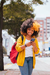 Positive African American female with curly hair and backpack smiling and browsing mobile phone while standing on city street by tree in daylight - ADSF45808