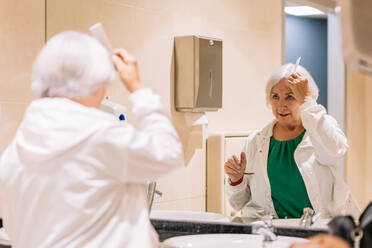 Elderly woman in white jacket standing near mirror in bathroom and brushing short gray hair - ADSF45817