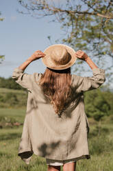 Back view of unrecognizable female in straw hat and casual wear relaxing on green field and admiring view of scenic landscape - ADSF45819