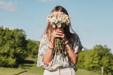 Faceless female in casual wear hiding behind fresh blooming bouquet of white wild daisy flowers on green field in summer - ADSF45821