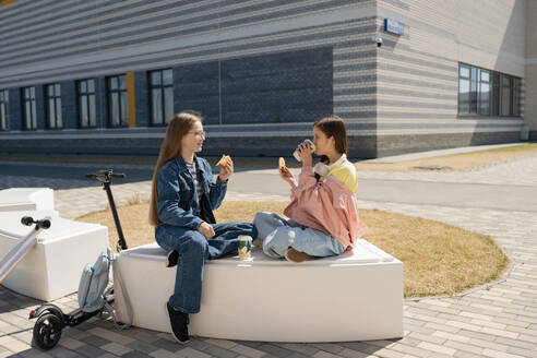 Side view of teenage girls in casual clothes having snacks and drinks sitting on bench near city building on sunny summer day - ADSF45822