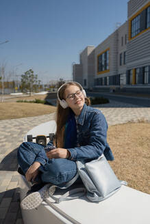 Trendy teenage girl in casual clothes sitting near bag while listening to music through headphones on white bench in city on summer day - ADSF45824