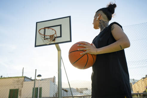 From below tattooed female basketball player with earphones looking away while holding ball near billboard on sports ground in daytime - ADSF45827