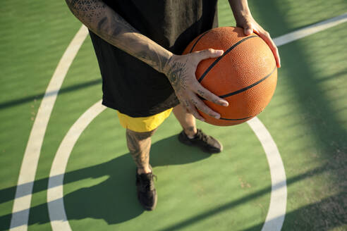 From above crop anonymous basketball player with tattoos holding ball while standing on sports ground - ADSF45828