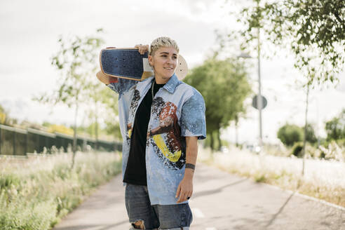 Positive shy young female in casual tomboy outfit holding with with the skateboard placed on shoulders on street and looking away - ADSF45829
