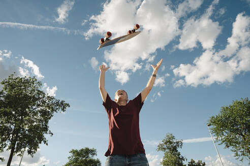 From below of young happy tomboy in casual clothes raising arms and tossing skateboard in sky while standing against blue cloudy sky - ADSF45830