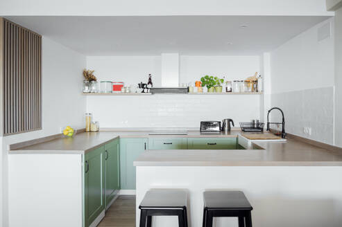 Interior design of minimal kitchen with marble top and cupboards with appliances and basin, white walls and shelf with bottles and stools near counter - ADSF45838