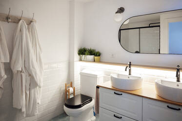 Minimal interior of bathroom with bathrobes on hanging stand and water sinks on wooden cabinet with shelves, mirror above and toilet bowl in corner - ADSF45839