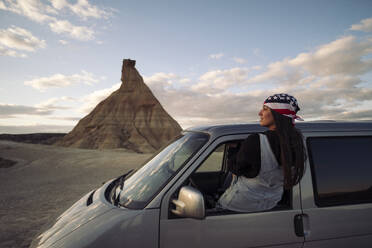 Back view of young female traveler in casual clothes with US scarf sitting on driver window of van while admiring beautiful dessert terrain in Navarra Spain - ADSF45852