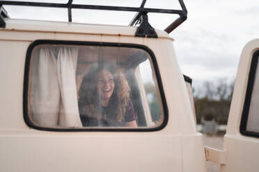 Through glass view of positive young female smiling and looking away while sitting near window with curtain in parked van with opened door - ADSF45859
