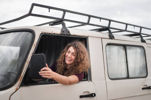 Cheerful young female with brown wavy hair touching rear view mirror and looking away while smiling and sitting in driver seat of parked van - ADSF45860