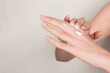 Close up of cropped anonymous female applying moisturizing cosmetic cream on manicured fingers with hand while standing in room with gray wall - ADSF45867