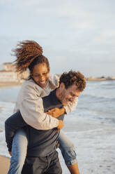 Smiling man giving piggyback ride to girlfriend at beach - JOSEF20355