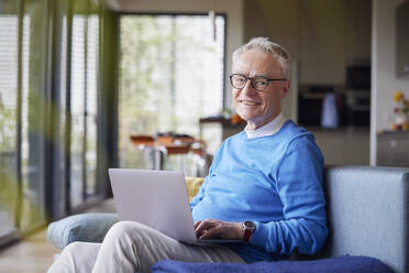 Smiling senior man using laptop on couch at home - RBF09132