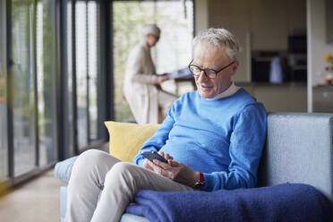 Senior man sitting on couch at home using mobile phone - RBF09137