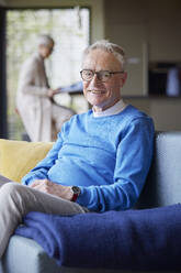 Portrait of smiling senior man sitting on couch at home with woman in background - RBF09139