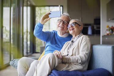 Happy senior couple sitting on couch at home taking selfie - RBF09140