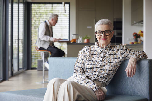 Porträt einer selbstbewussten älteren Frau, die zu Hause auf der Couch sitzt, mit einem Mann im Hintergrund - RBF09141