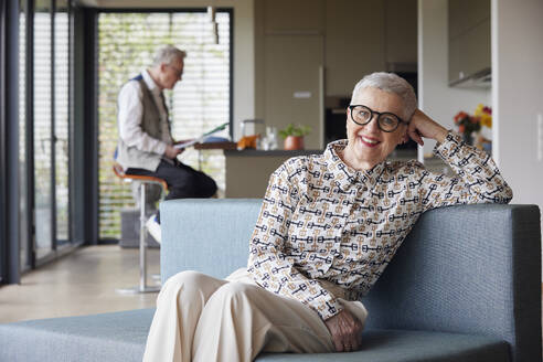 Porträt einer lächelnden älteren Frau, die zu Hause auf der Couch sitzt, mit einem Mann im Hintergrund - RBF09142