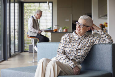 Smiling senior woman sitting on couch at home with man in background - RBF09144