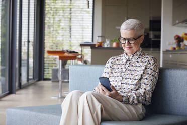 Senior woman sitting on couch at home using mobile phone - RBF09146