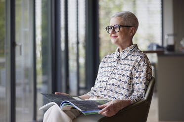 Senior woman sitting in armchair at home with magazine - RBF09185