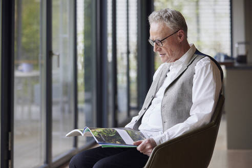 Senior man sitting in armchair at home reading magazine - RBF09189