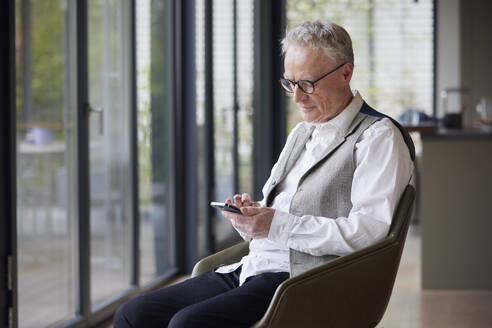 Senior man sitting on chair at home using mobile phone - RBF09190