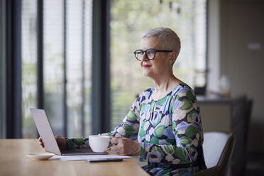 Senior woman sitting at table at home with laptop and coffee cup - RBF09199