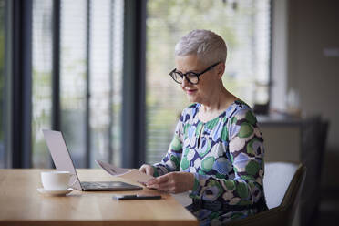 Senior woman with document and laptop at table at home - RBF09200
