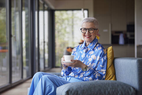 Smiling senior woman sitting on couch at home with cup of coffee - RBF09220