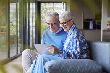 Happy senior couple sitting on couch at home using tablet PC - RBF09228